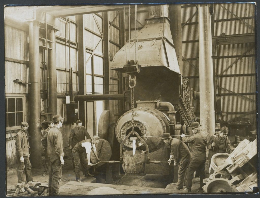 Sepia photograph of a factory scene. There is a large bucket hanging from a chain and pulley assembly, with two men holding the handles either side of the bucket. A third man guides molten metal into the bucket from a large furnace.